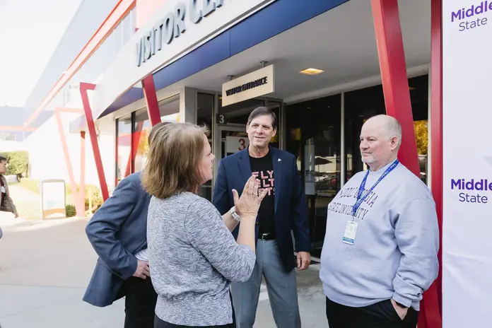 Four people talking outside a Visitor Center entrance with red beams; one wears a ‘Fly Delta Jets’ shirt, another a Middle State sweatshirt.