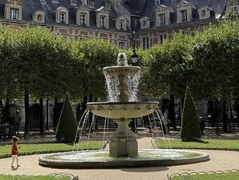 Stone fountain in a leafy Paris square, framed by manicured trees and historic red-brick buildings with slate roofs.