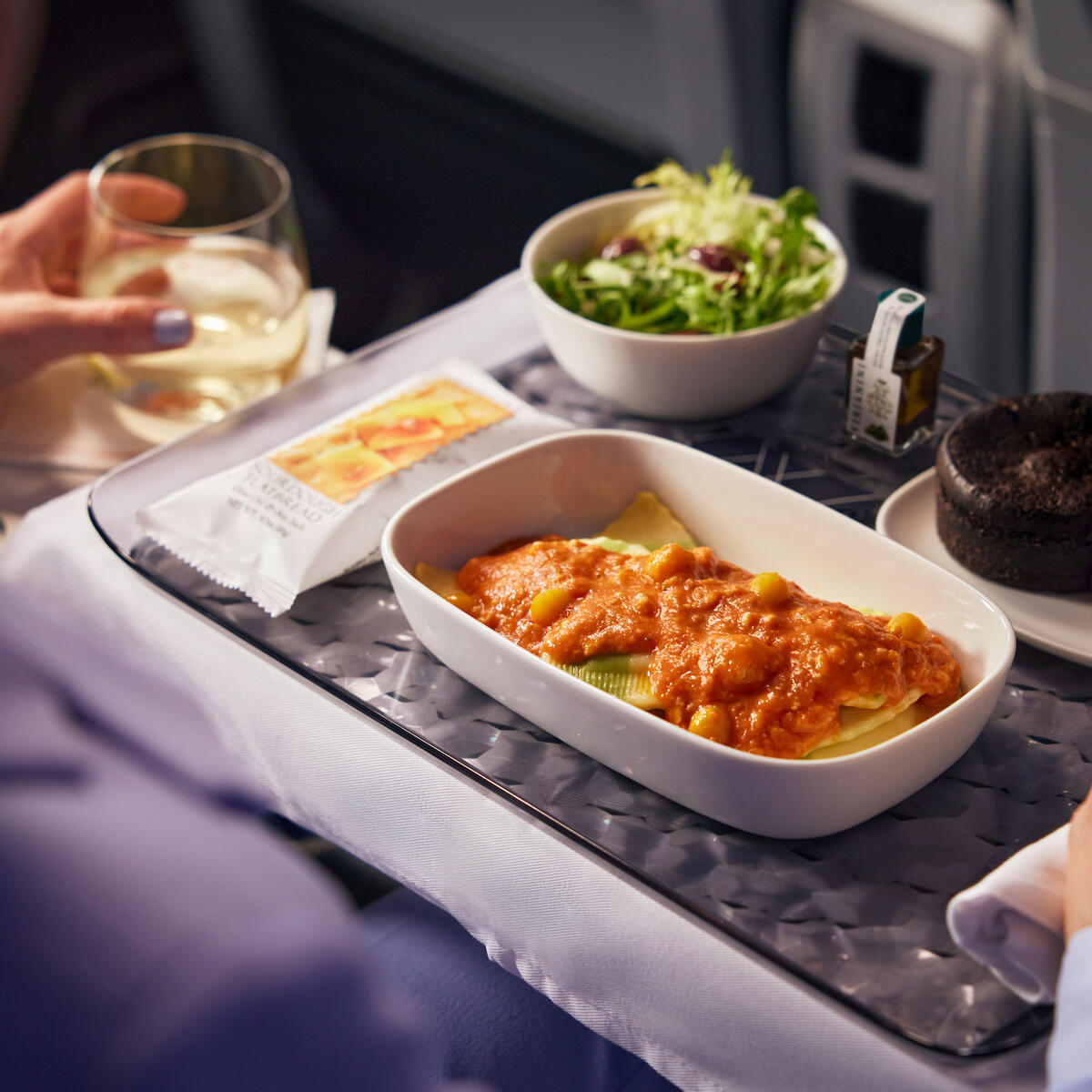 A meal served on Delta Premium Select: Spinach and Ricotta Tortellini, tossed in red pepper mascarpone sauce, accompanied by roasted vegetables, crumbled feta, and nut-free basil pesto, with a decadent dessert: Chocolate Brownie Passion Fruit Bar  