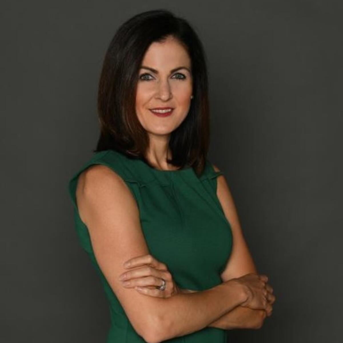 Studio portrait of a person with shoulder-length dark hair, wearing a sleeveless green dress and standing with arms crossed against a plain gray background.