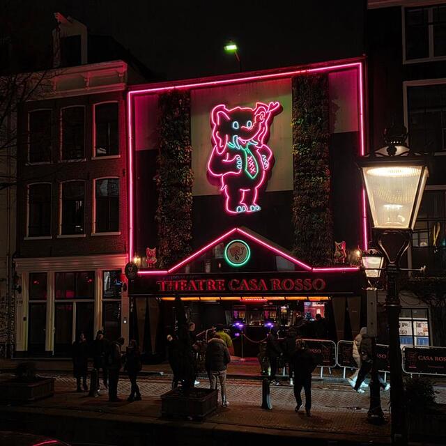 Nighttime view of a building with bright pink neon lights and a large neon elephant sign above the entrance to Theatre Casa Rosso, with people walking along the canal in front.