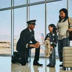 Delta pilot kneeling beside a young child holding a stuffed animal at an airport gate, while an adult stands nearby with rolling luggage; airplane visible through large windows in the background.