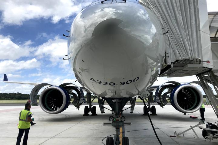 Nose of A220-300 parked on tarmac with stair truck and employees