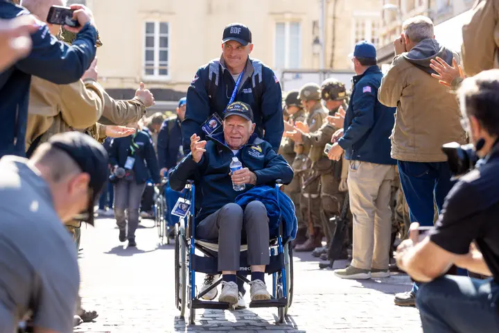 Army veteran Tom Lacey and his caretaker, Air Force veteran and Atlanta-based 717 Capt. Brandon Williams, wave to fans at a parade in Carentan.