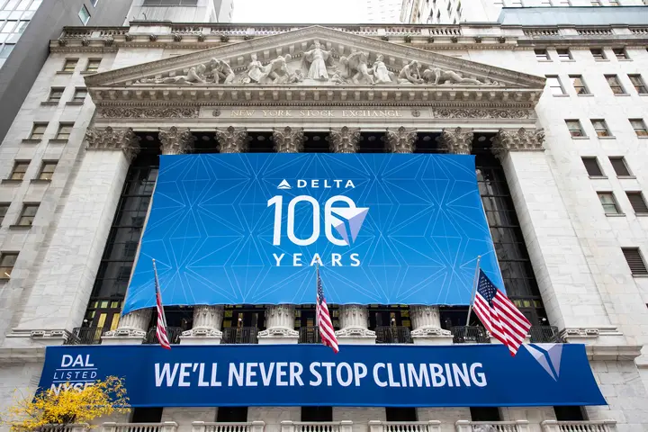 Exterior shot of NYSE building with a large, rectangular banner displaying Delta's centennial logo and a smaller banner below that reads We'll Never Stop Climbing