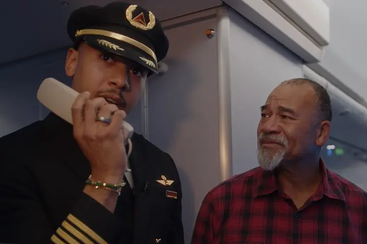 Airline pilot in uniform stands inside an aircraft cabin near the galley, holding an intercom handset. Another person wearing a red plaid shirt is standing beside the pilot.