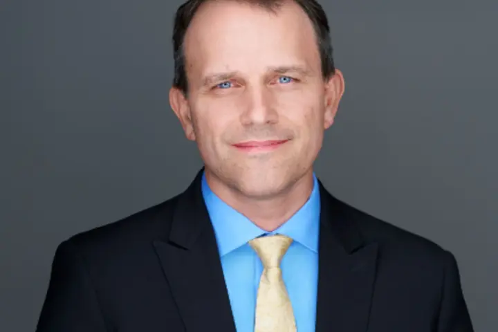 Headshot of Matt Schrag, wearing a dark suit jacket, light blue dress shirt and light yellow tie, posed against a plain gray background.