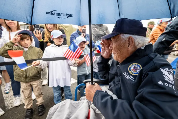 An older person in a veteran’s jacket sits under an umbrella while children hold small American flags nearby during an outdoor commemorative event.