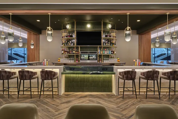 Front view of lounge bar with high‑back stools, illuminated shelving, pendant lights and wood‑pattern flooring