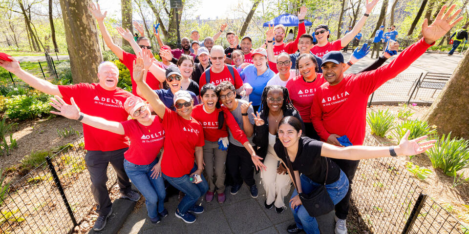 A large group of Delta volunteers wearing red “Delta in the Community” shirts stand together in a park with arms raised, gathered along a paved path surrounded by trees.