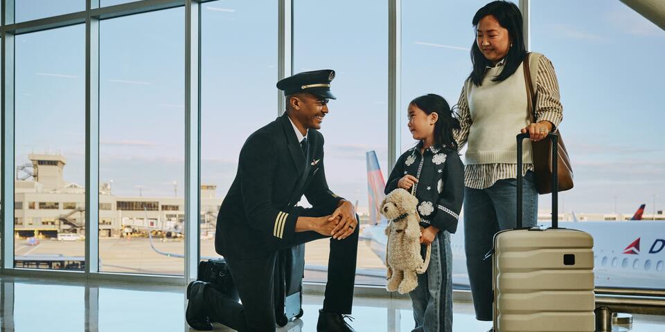 Delta pilot kneeling beside a young child holding a stuffed animal at an airport gate, while an adult stands nearby with rolling luggage; airplane visible through large windows in the background.