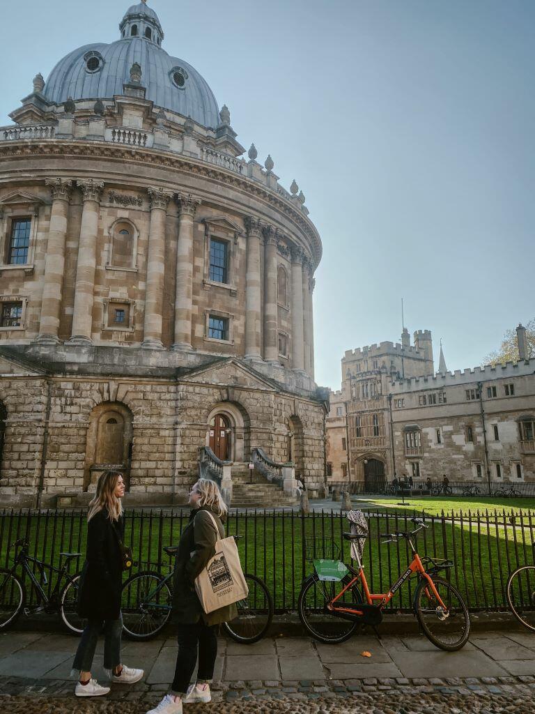 Two people stand in front of the Radcliffe Camera building at the University of Oxford.
