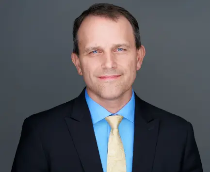 Headshot of Matt Schrag, wearing a dark suit jacket, light blue dress shirt and light yellow tie, posed against a plain gray background.