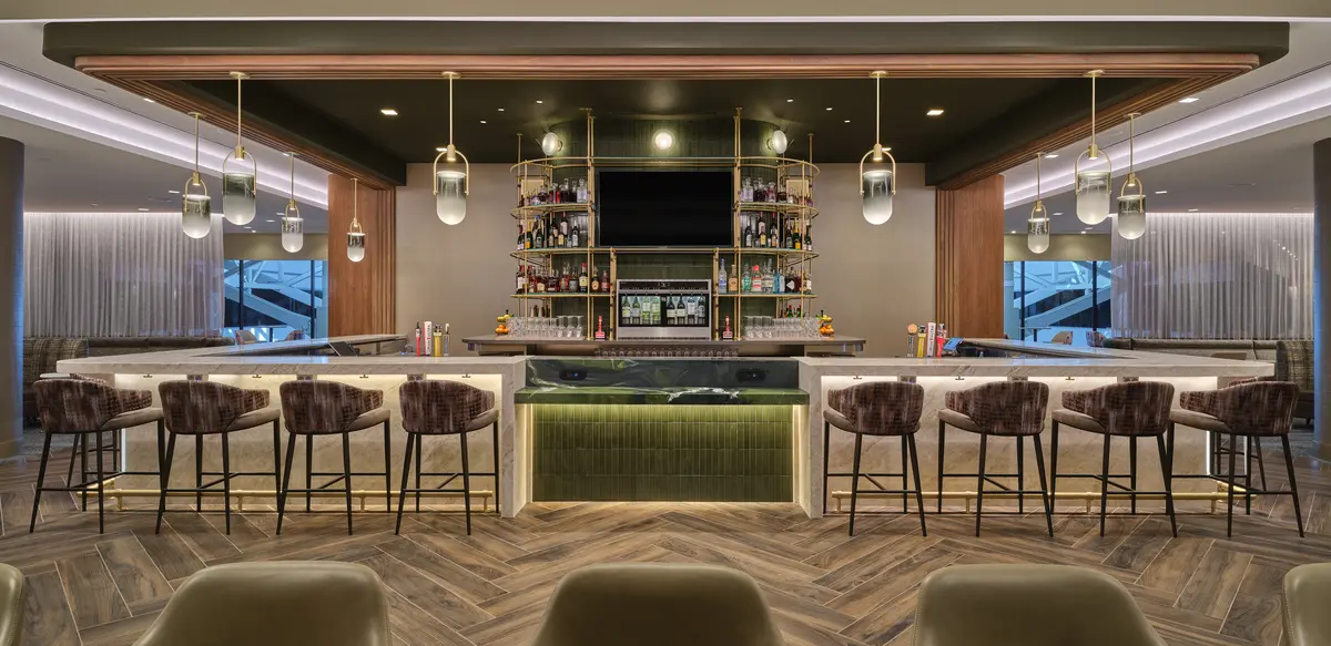 Front view of lounge bar with high‑back stools, illuminated shelving, pendant lights and wood‑pattern flooring