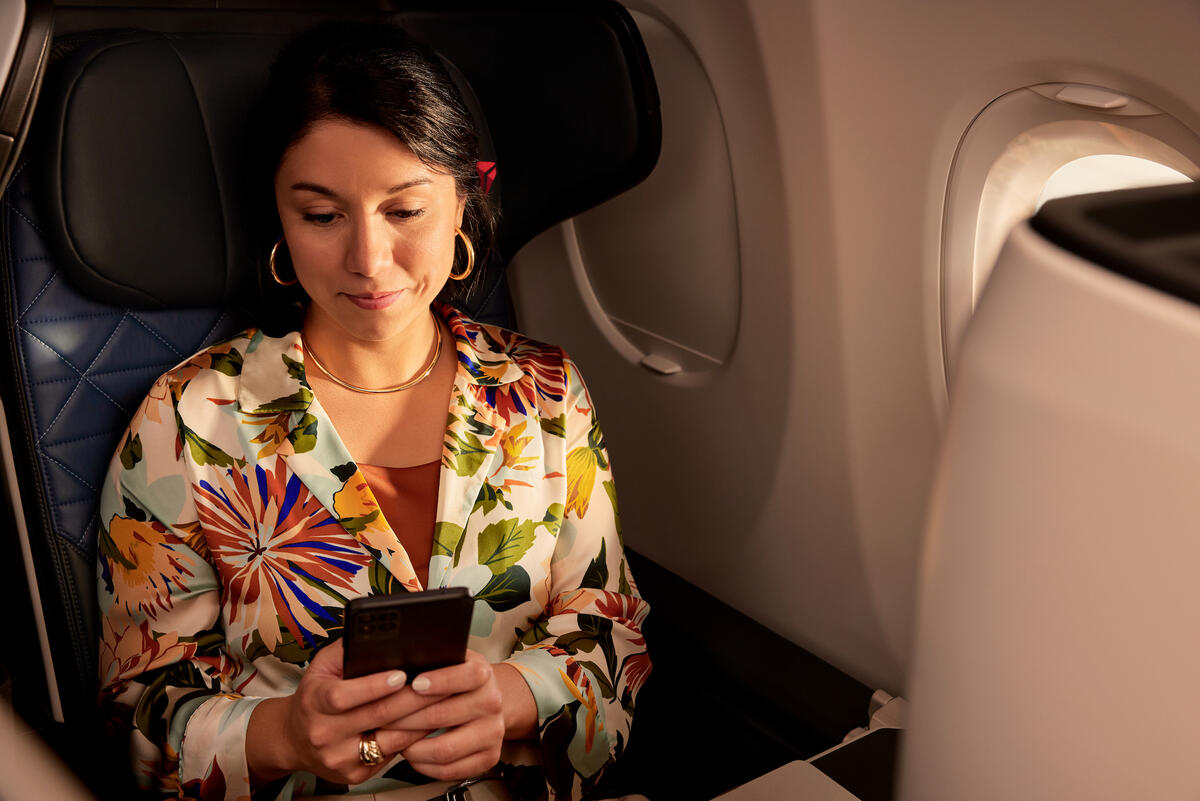 Passenger seated by an airplane window holds a smartphone while sitting in a premium cabin seat, with soft lighting and aircraft interior visible.